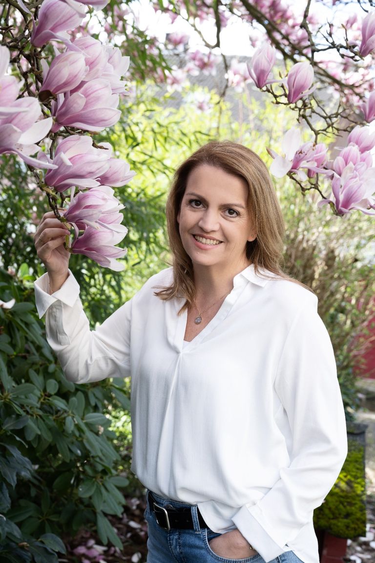 Adina Ortjohann steht in weißer Bluse und Jeans unter einem blühenden Magnolienbaum und lächelt