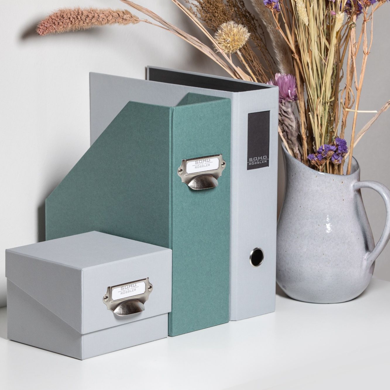 Büro-Organisationsprodukte S.O.H.O. by Rössler: Stehsammler, Ordner und Box in Grau und Grün neben einer Vase mit Trockenblumen